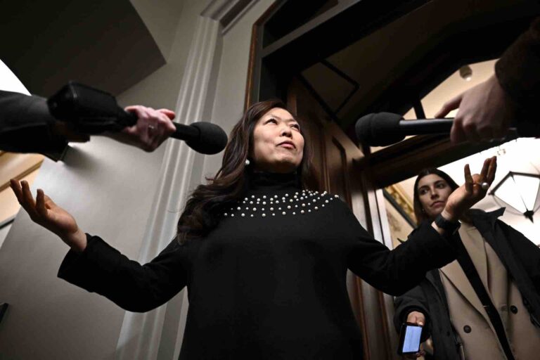 Trade Minister Mary Ng wears a black sweater with white pearls. Her arms are outstretched as two mics point toward her. The photo is taken from a low angle, looking up at Ng.