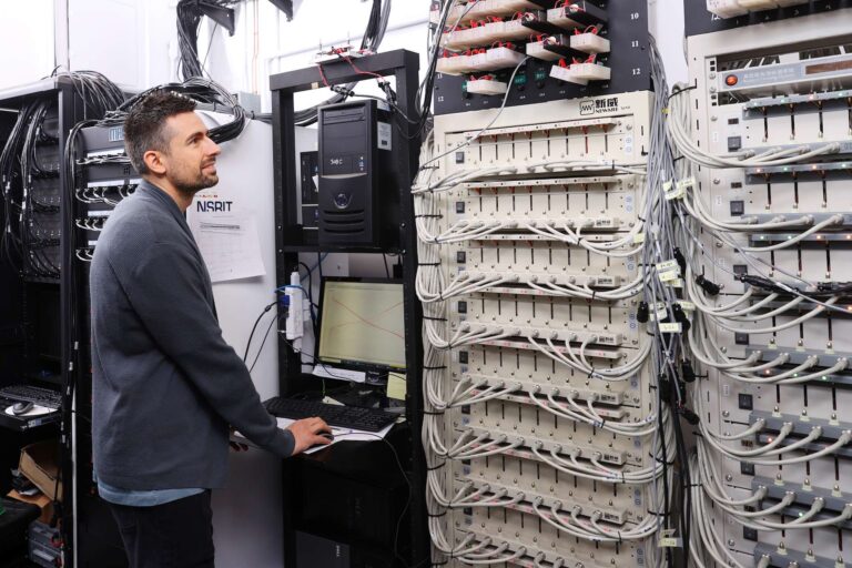 Michael Metzger, pictured with short, dark hair wearing a grey sweater, types on a computer while looking up at a wall covered with wires at one of Dalhousie University's current battery testing labs in Halifax, Nova Scotia.