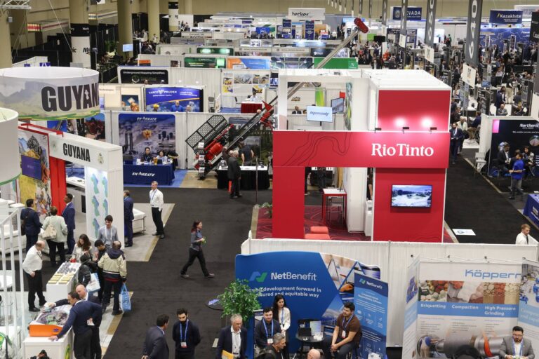 Dozens of booths in a conference centre at the Prospectors & Developers Association of Canada’s annual convention, with two large white signs saying “Guyana” on the left, a large red booth for RioTinto on the right and a smaller blue booth for NetBenefit at the bottom. People are seen talking at the booths and a tall piece of mining equipment is on display farther back.