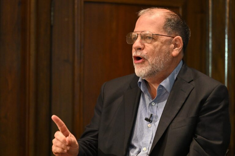 Tyler Cowen sits facing the left on the right side of this image. He wears ‘70s style eyeglasses and a black blazer. His finger is pointing to the left.