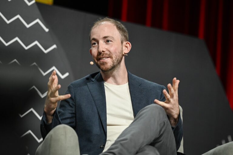 Wealthsimple CEO Michael Katchen sitting in a chair, gesturing with both hands. He is wearing a white T-shirt with a blue blazer and has a microphone attached to his ear.