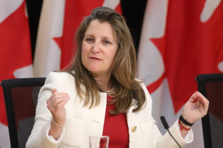 Finance Minister Chrystia Freeland sits in a chair at a news conference. She wears a white blazer and a red top. Her hands are gesturing as she speaks.