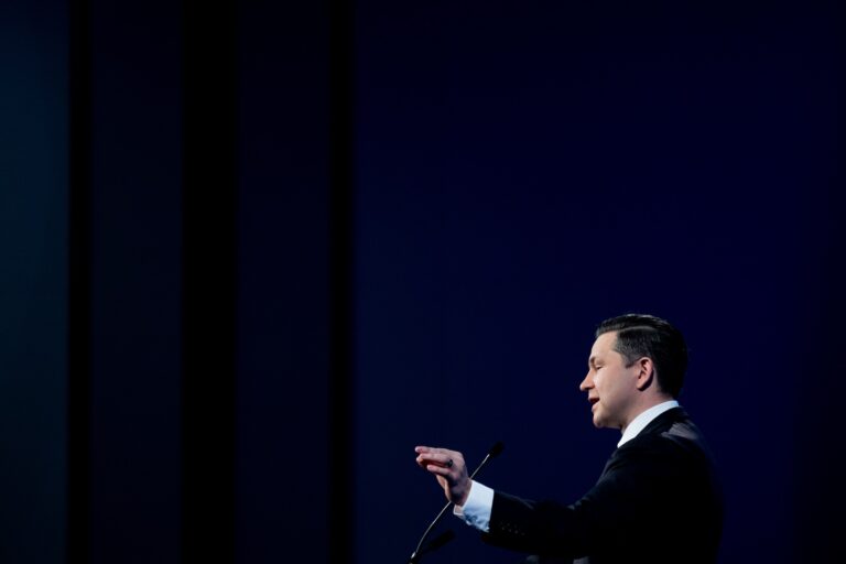 Conservative Party Leader Pierre Poilievre speaking into a microphone and gesturing with his left hand on a dark blue background.