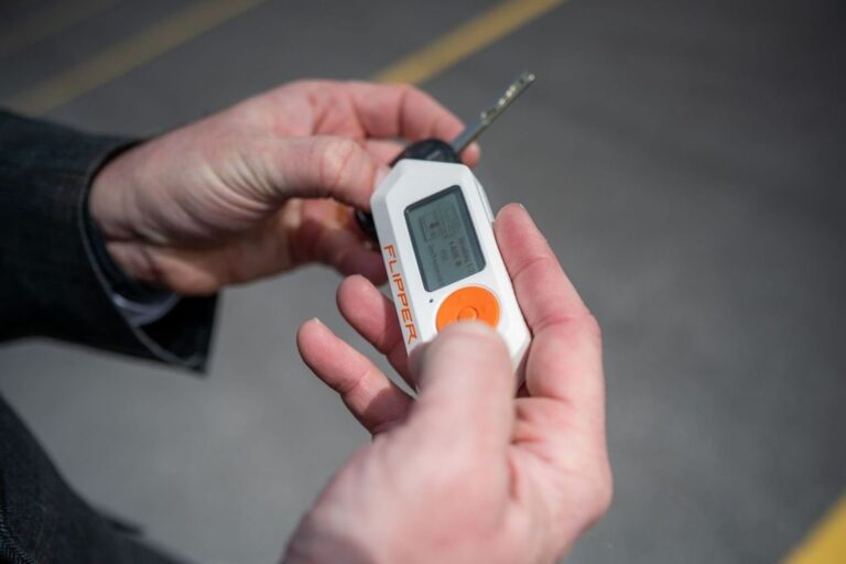 Two hands holding a car key, and a small device with a tiny screen, a button and text that says ‘FLIPPER’ in orange colour.
