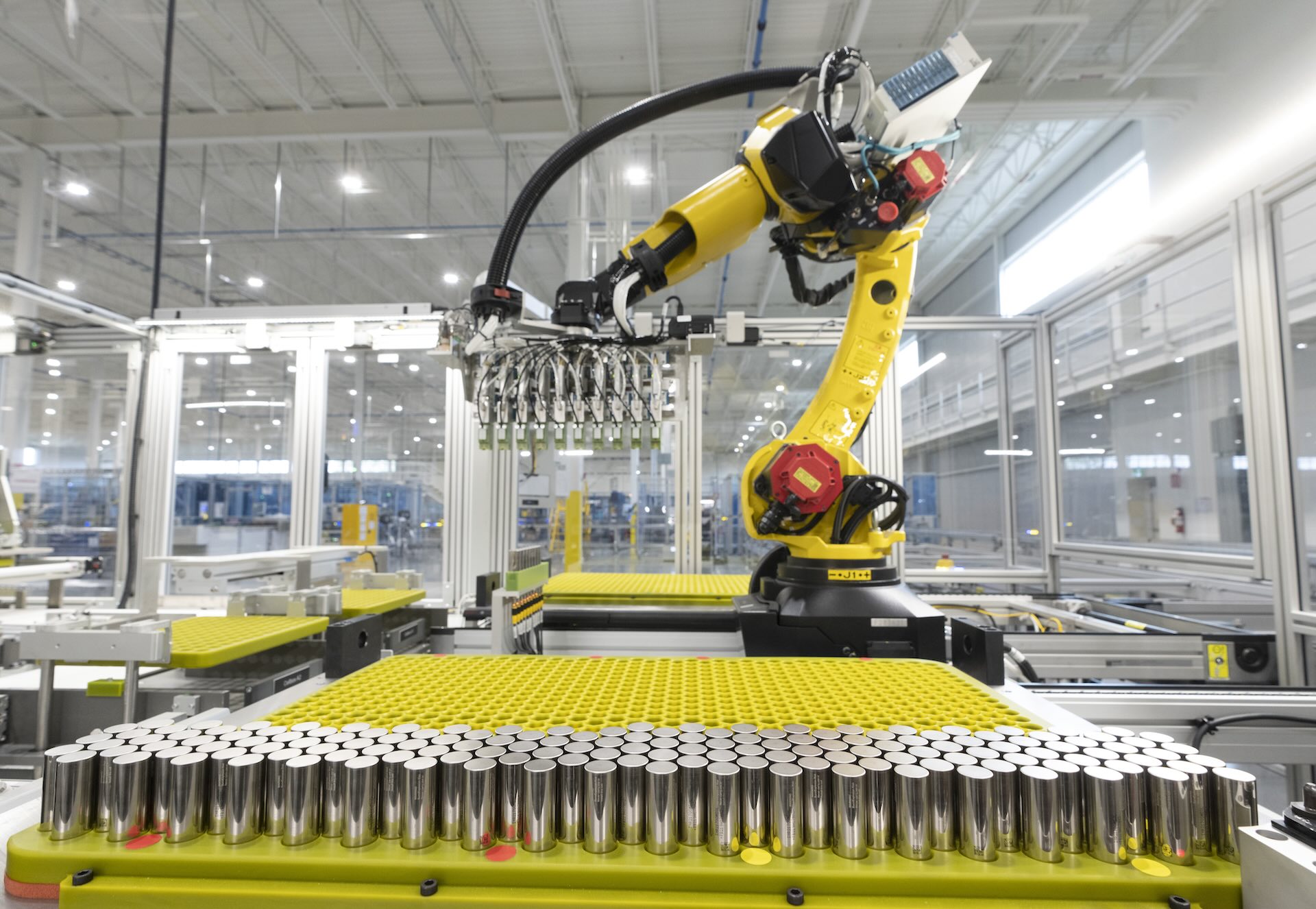 A robotic arm operating a machine in a factory with a stack of lithium-ion batteries below it.