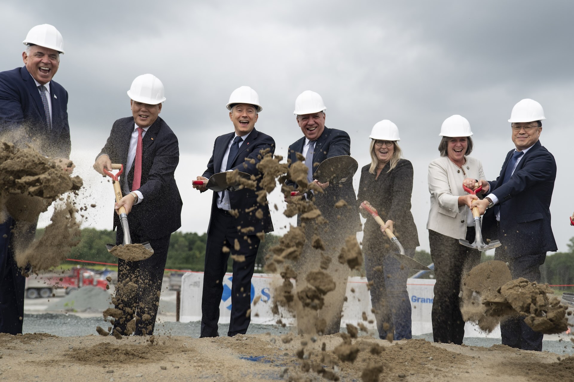 Federal and Quebec innovation ministers, along with Ford Canada and SK On executives, toss dirt with their shovels toward the camera during a groundbreaking ceremony at a construction site.
