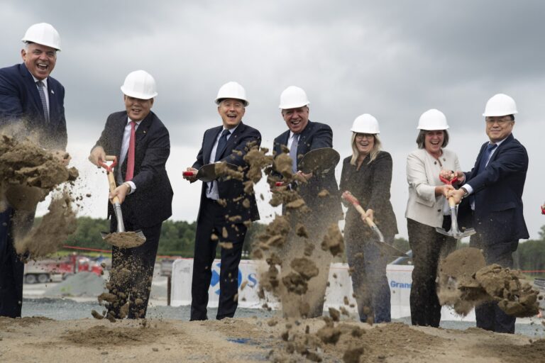 Seven politicians and EV business executives, wearing white hard hats, throw dirt into the air with shovels while laughing and smiling at a ground-breaking ceremony. A construction zone is behind them.