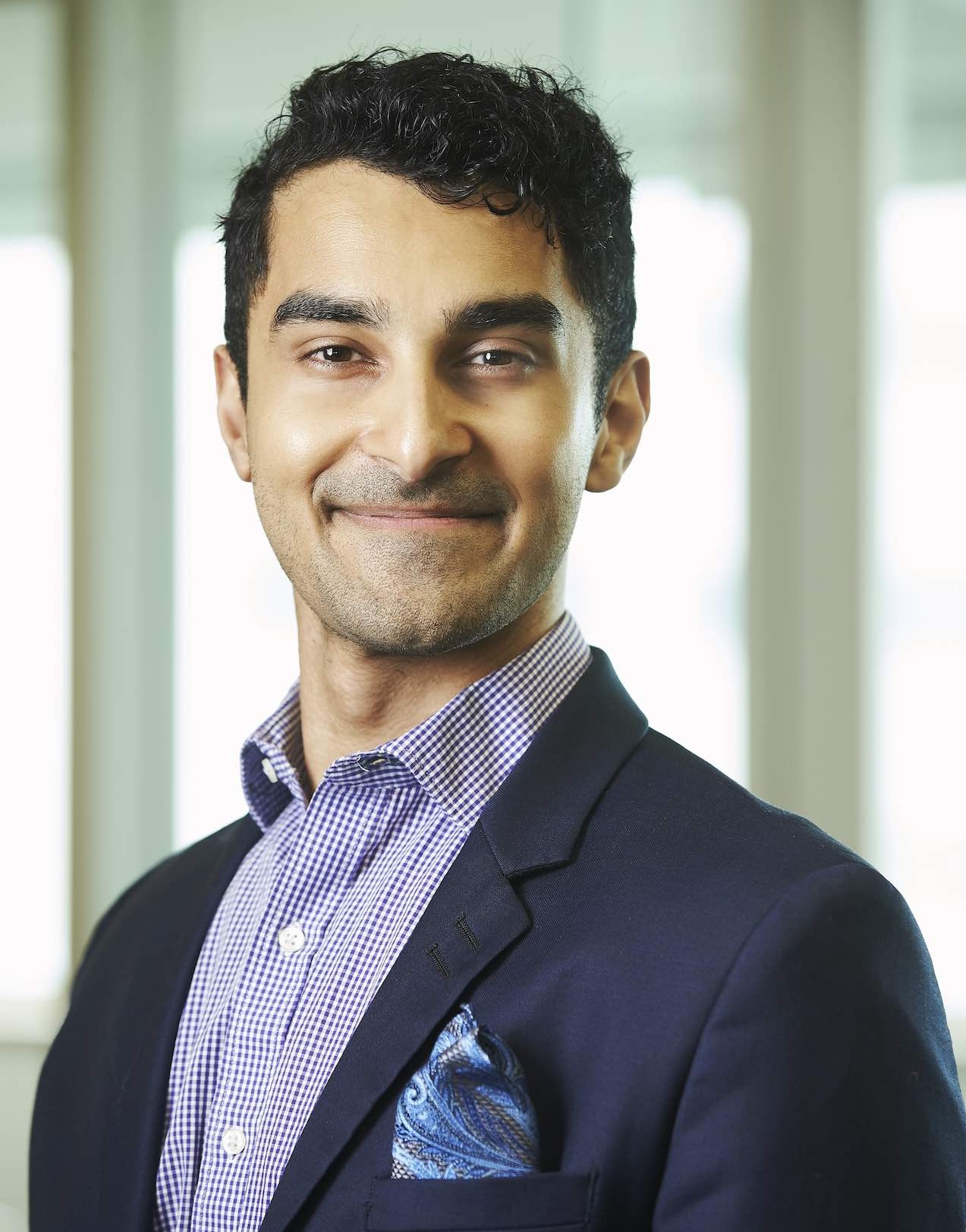 Saumik Biswas smiles toward the camera in a headshot. He wears a navy blazer and a purple checkered shirt.
