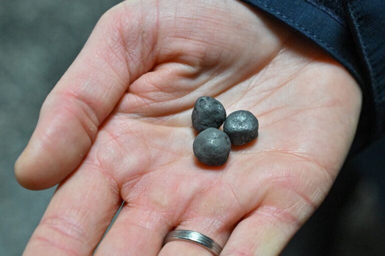 An open palm holds three small spheres of iron. The hand also wears a silver band on its ring finger.