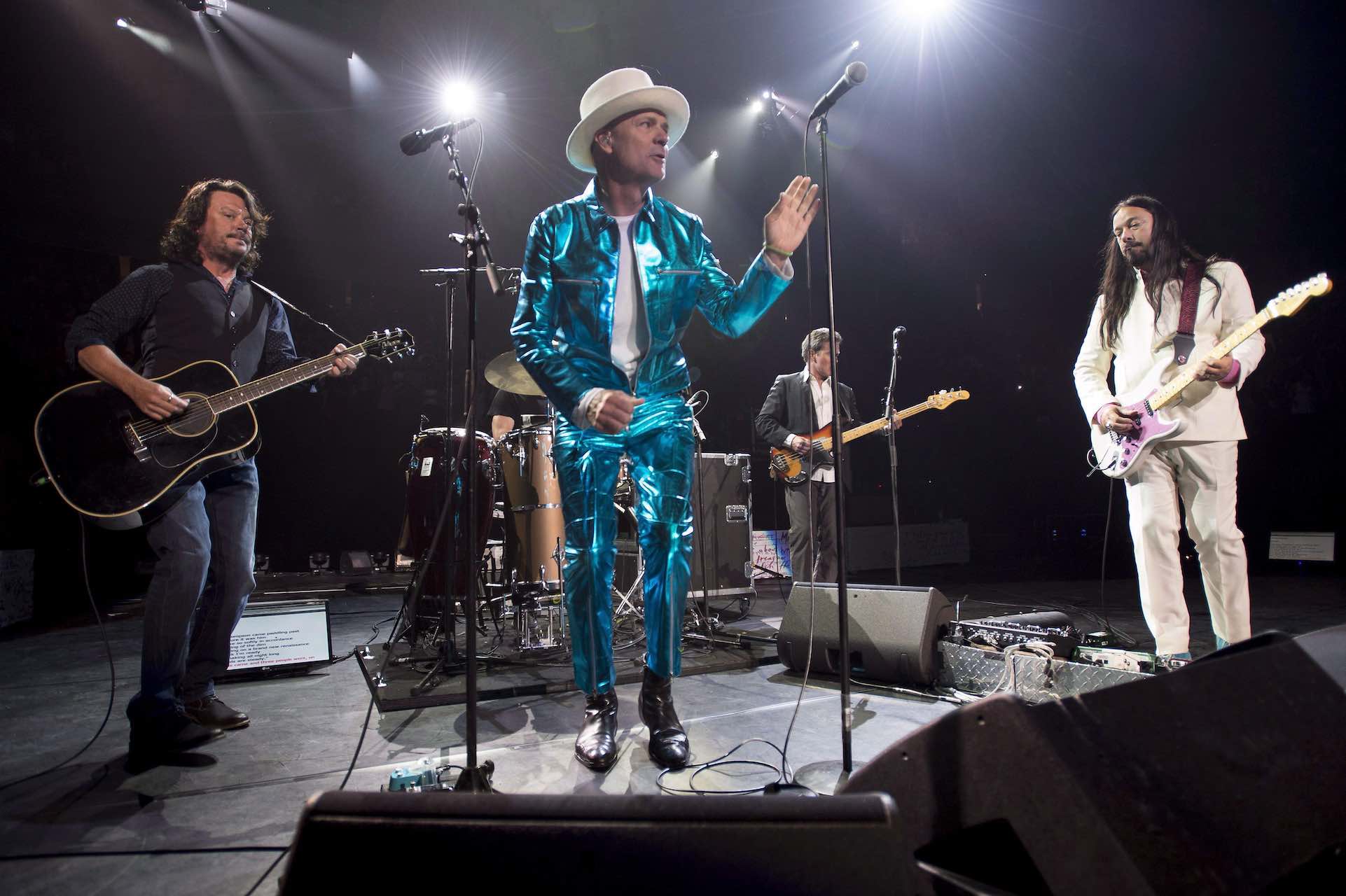 Members of the Tragically hip stand in five different spots on stage. Frontman Gord Downie wears a metallic blue suit and a white hat, his left hand gestures toward a mic in a stand.