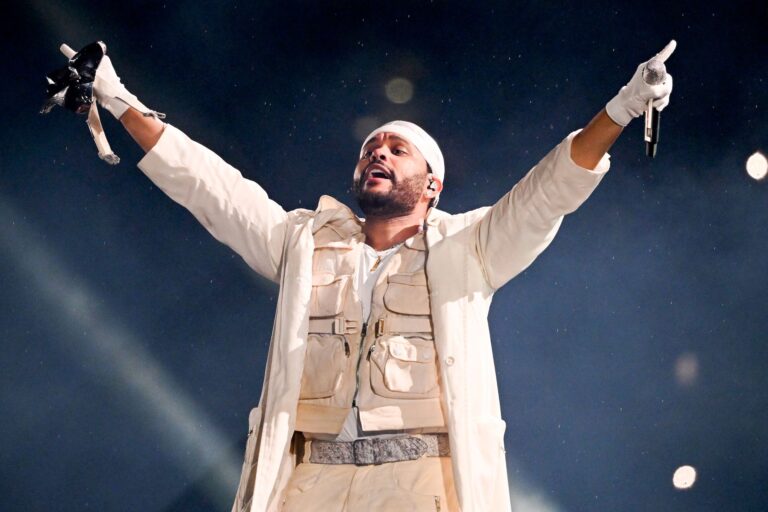 Musician The Weeknd performs in a white outfit. Both his hands are raised outwards and he holds a microphone in his left hand.