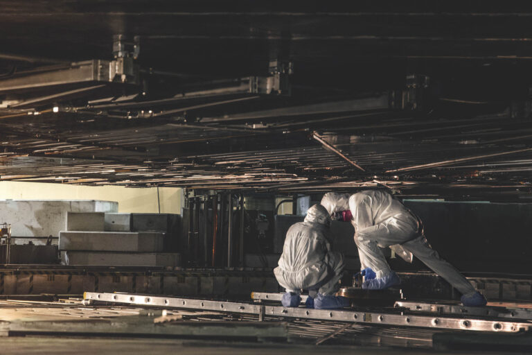 Two workers wearing white coveralls with hoods, masks and blue booties crouch while performing work in the narrow confines of a dark, metal-lined chamber.