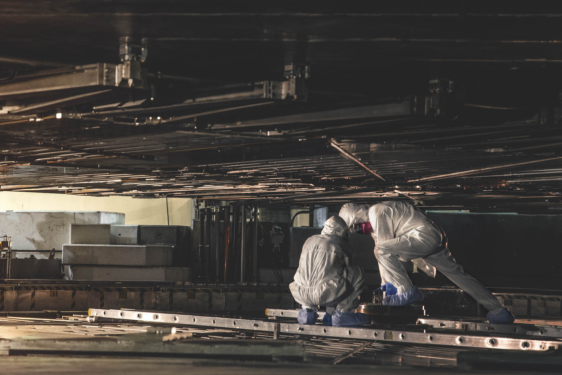 Two workers wearing white coveralls with hoods, masks and blue booties crouch while performing work in the narrow confines of a dark, metal-lined chamber.