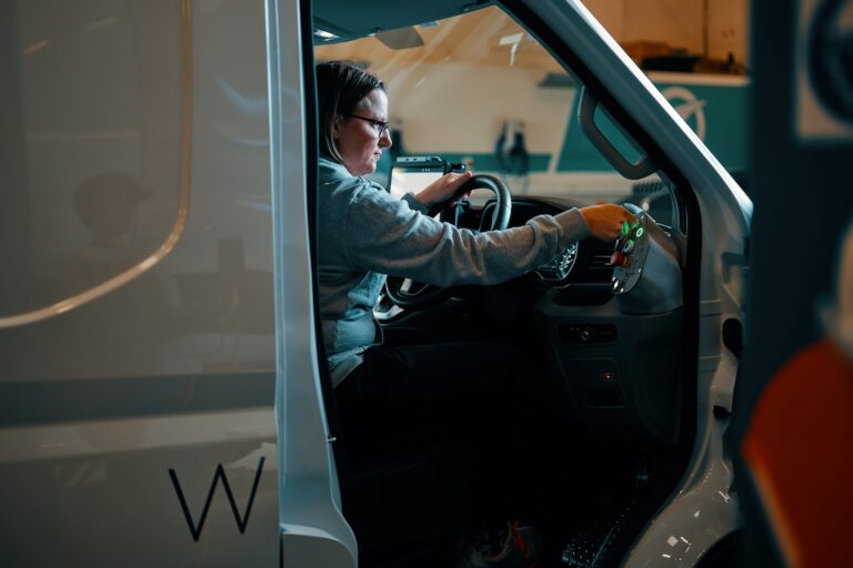 A person with shoulder-length hair and glasses sits in a white van with the door open, with one hand on the steering wheel and the other hand on a console of colourful buttons. The van has a “W” on the side.