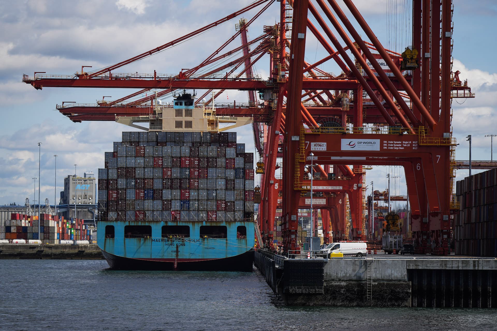 A baby-blue hulled ship with a load of sea containers is docked beneath a row of towering red cranes.