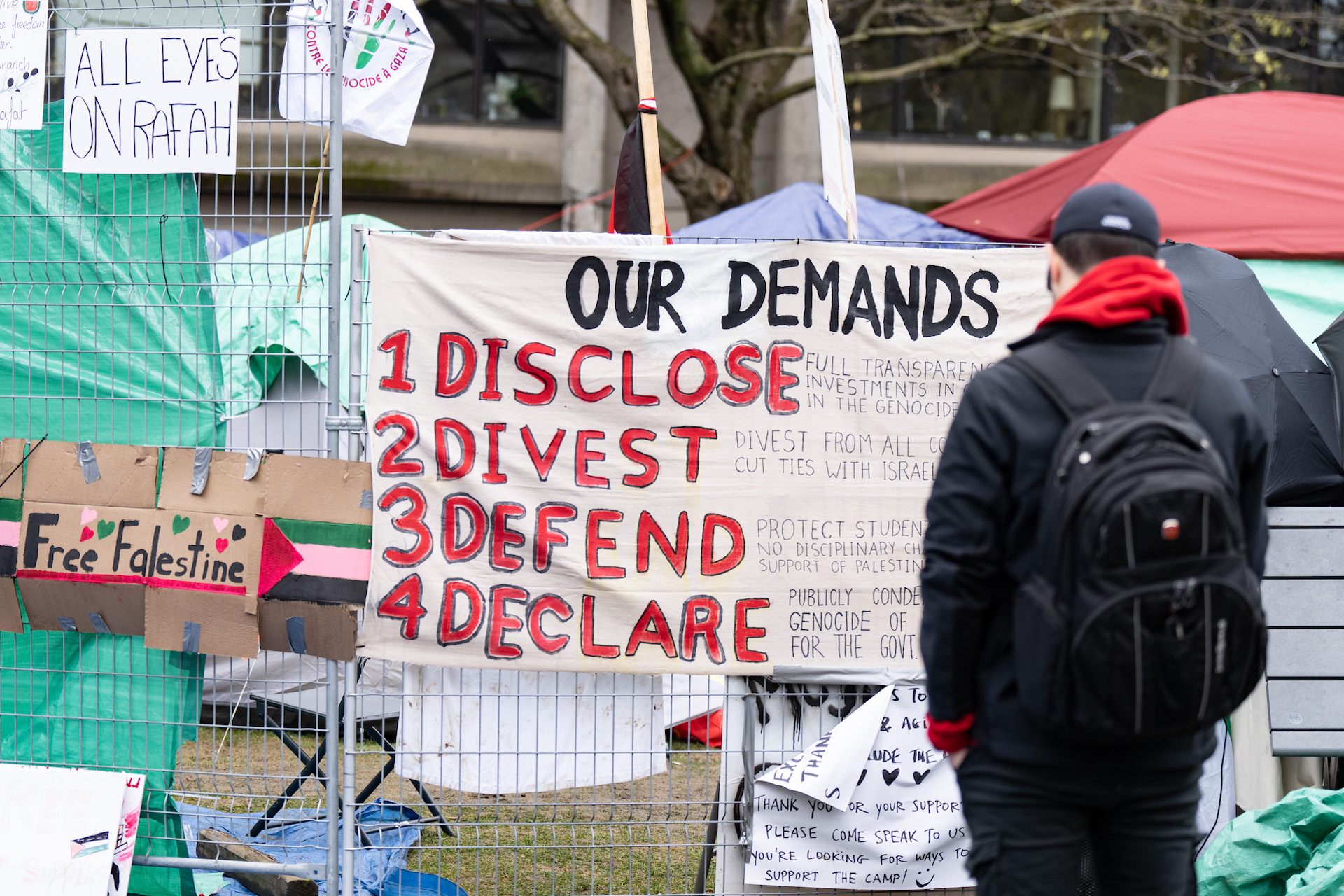 A black-clad man with his back to the camera stands before a poster hung on a section of wire fencing around a tent encampment. The poster reads "Our demands: disclose, divest, defend, declare."