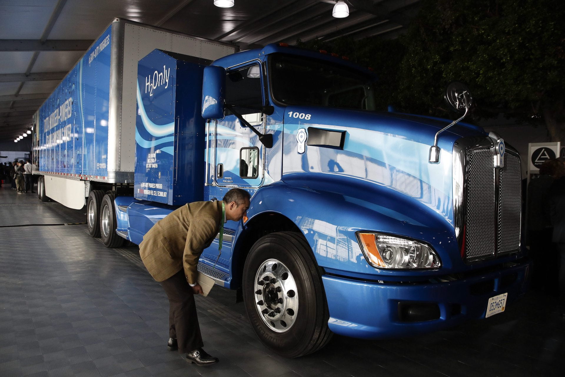 A man in a beige suit leans in for a close look at a shiny blue tractor-trailer unit emblazoned with the label "H2Only."