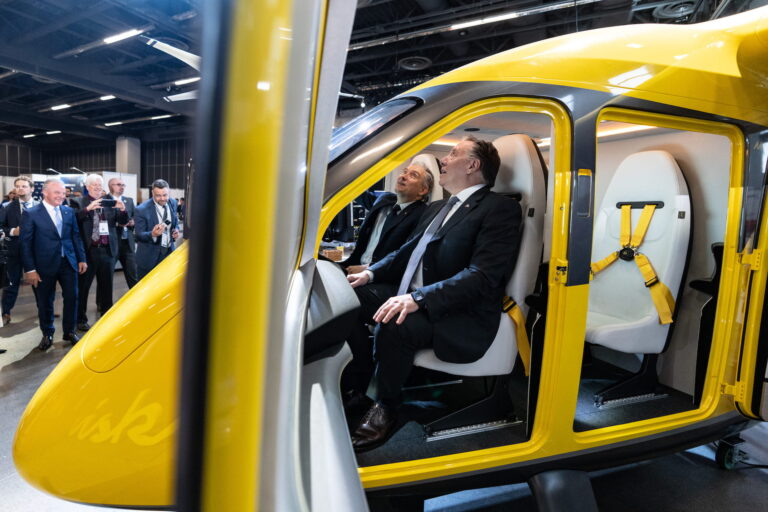 Legault and Champagne, both wearing dark suits, sit smiling in the four-seat cabin of a yellow, helicopter-like aircraft.