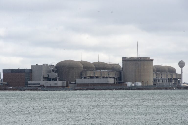 A cluster of large, concrete industrial-style buildings is visible across the waters of Lake Ontario. The complex includes seven cylindrical structures with dome-shaped tops.
