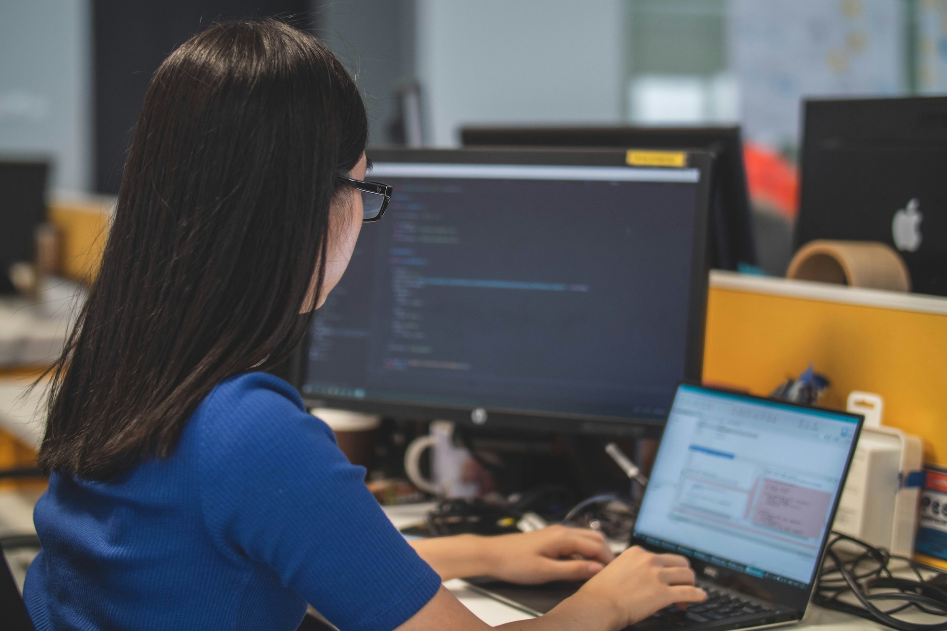 A person in glasses and a blue top is sitting and typing on a laptop in an office. A desktop screen next to the laptop displays some blurred-out coding work.