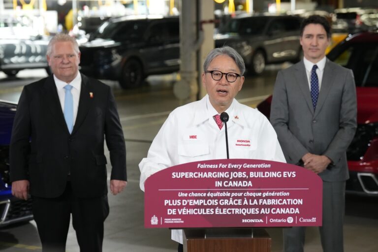 Prime Minister Justin Trudeau and Ontario Premier Dog Ford look on as Honda executive Toshihiro Mibe speaks in a white Honda lab coat at a podium that says, “Supercharging jobs, building EVs in Canada.”