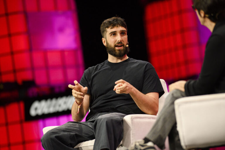 Cohere CEO Aidan Gomez sitting and speaking on stage, with a microphone attached to his ear. Background features illuminated panels and a large sign reading "COLLISION.”