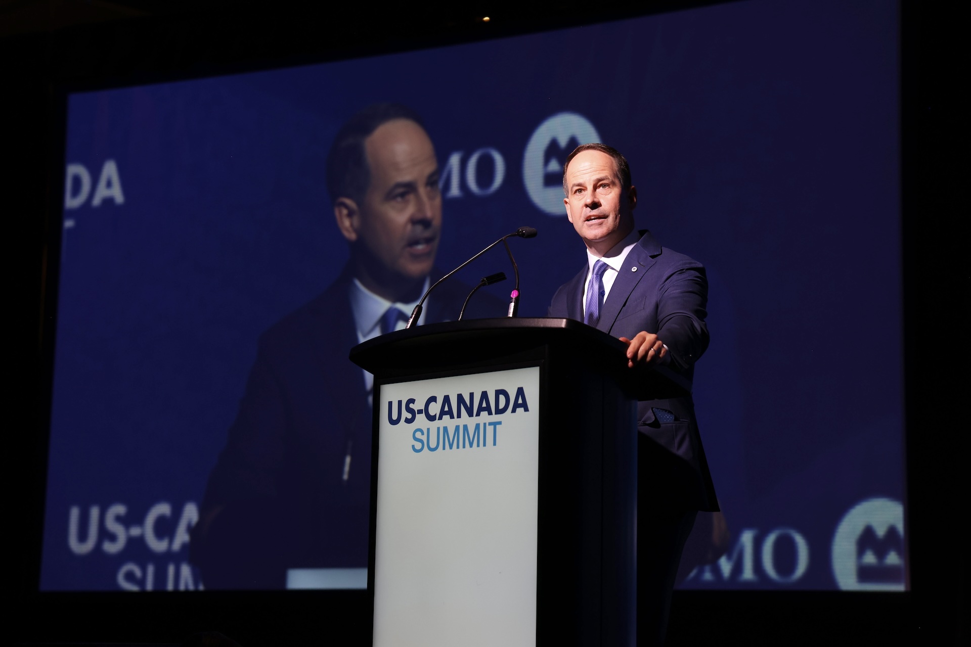 Bank of Montreal CEO Darryl White standing at a podium, showing the U.S.-Canada Summit logo, in front of a projector screen.