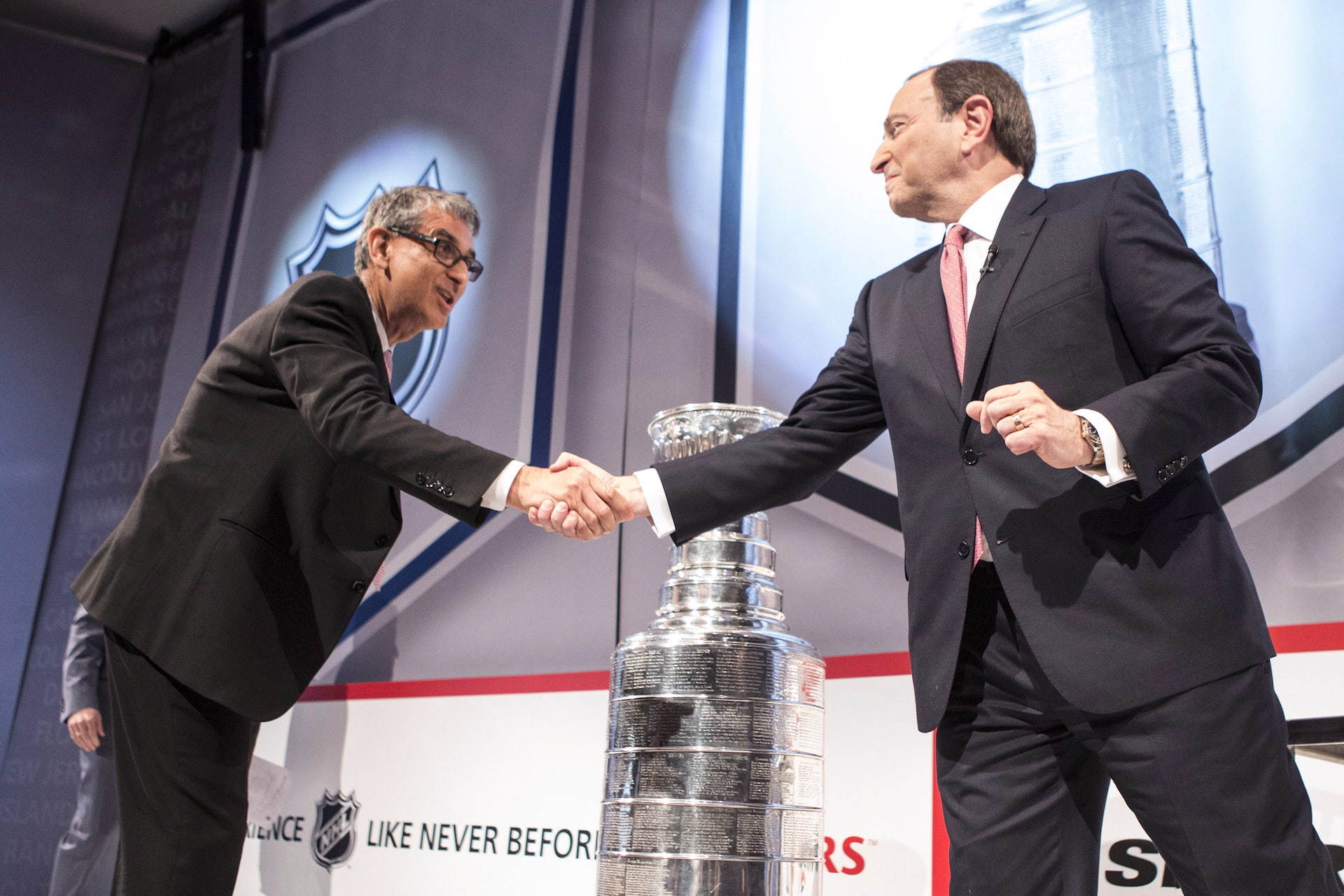 Nadir Mohamed bends forward to shake the hand of Gary Bettman at a photo-op, with the Stanley Cup displayed in the background. Both men are wearing dark suits.