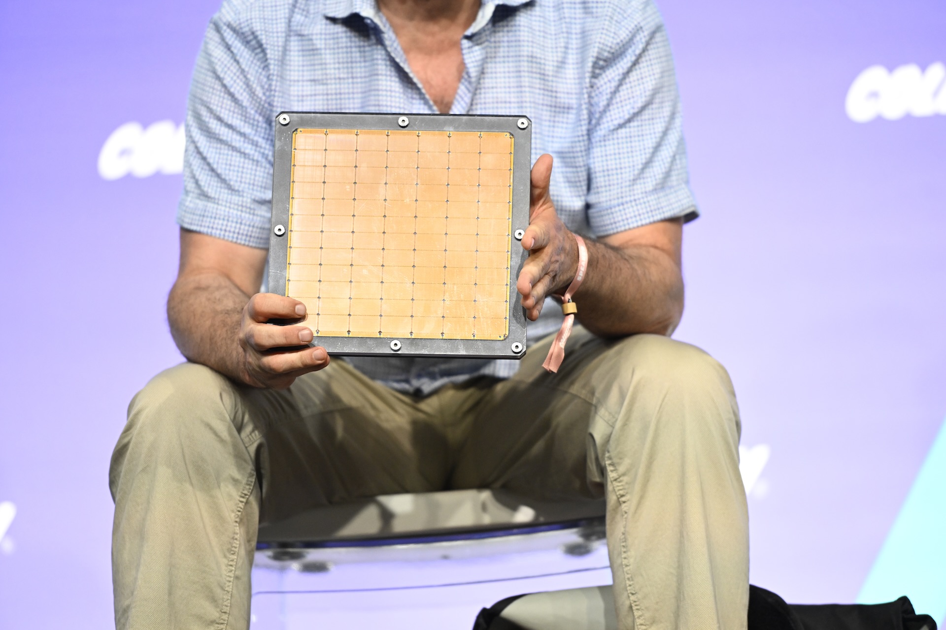 A person in a checkered shirt and khaki pants is seated holding up a square electronic component with a grid pattern against a purple backdrop.