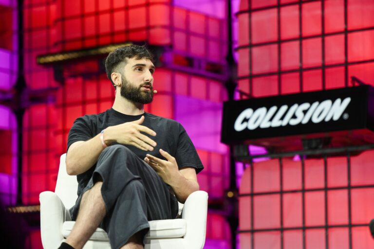 Cohere CEO Aidan Gomez sitting and speaking on stage, with a microphone attached to his ear. Background features illuminated panels and a large sign reading "COLLISION.”