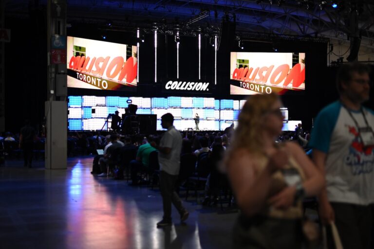 A large room with people attending the Collision event in Toronto. The stage has screens displaying "Collision" and "Toronto."