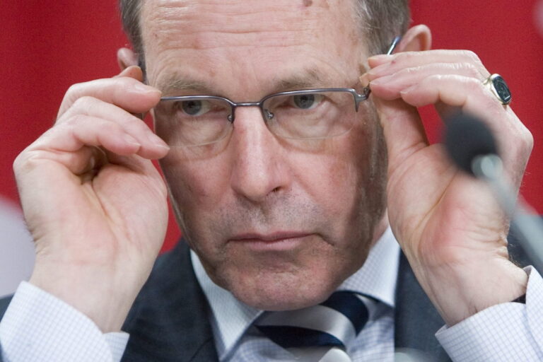 A close-up shot of former Bank of Canada governor David Dodge, clean-shaven and balding, against a backdrop of Canadian flags. Dodge is adjusting his glasses.