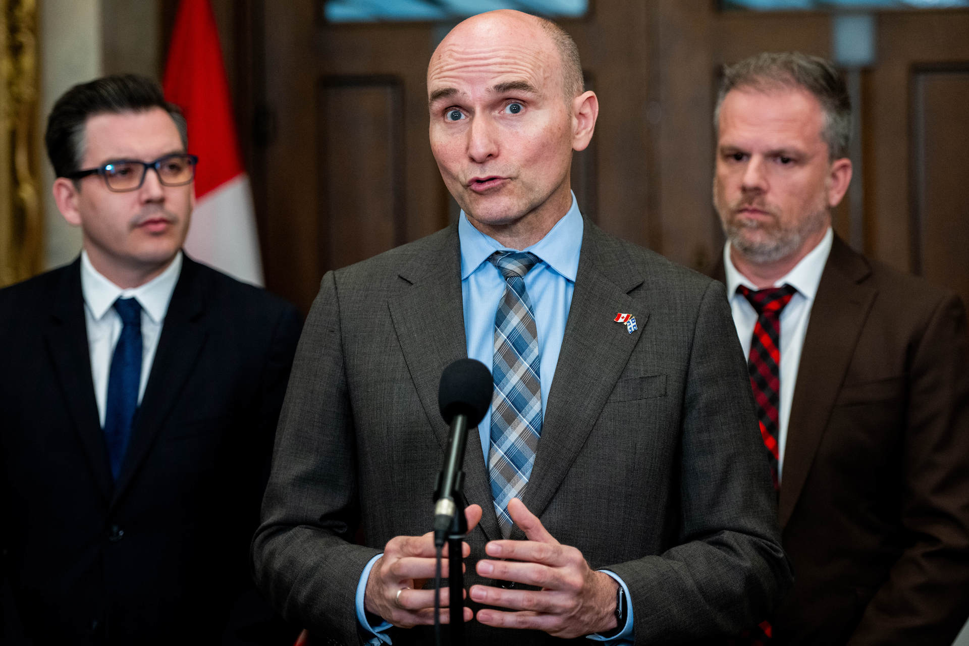 Jean-Yves Duclos, in a brown suit jacket with a blue shirt and tie, stands behind a uni-mic in the Commons foyer flanked by fellow Liberal MPs. Duclos's head is shaven near-bald, and he is wearing a lapel pin featuring the Canadian and Quebec flags.