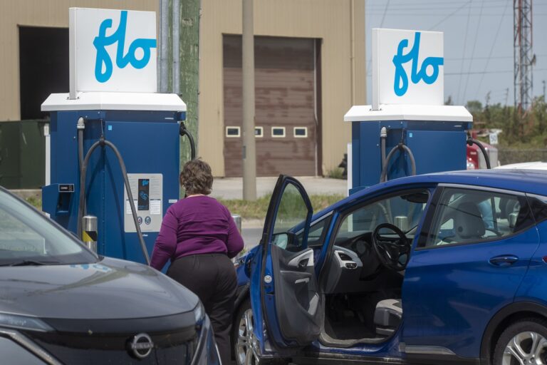Two cars parked at a station where two charging units bear the logo "Flo." The driver-side door of one small, blue car is open, and a woman is approaching one of the charging ports.