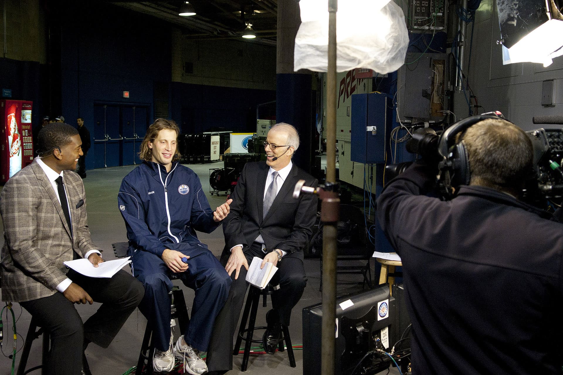 Tom Gilbert, a long-haired player wearing an Edmonton Oilers warmup suit, sits on a stool under TV lights between two besuited hosts. A TV camera operator appears in the right-hand foreground.
