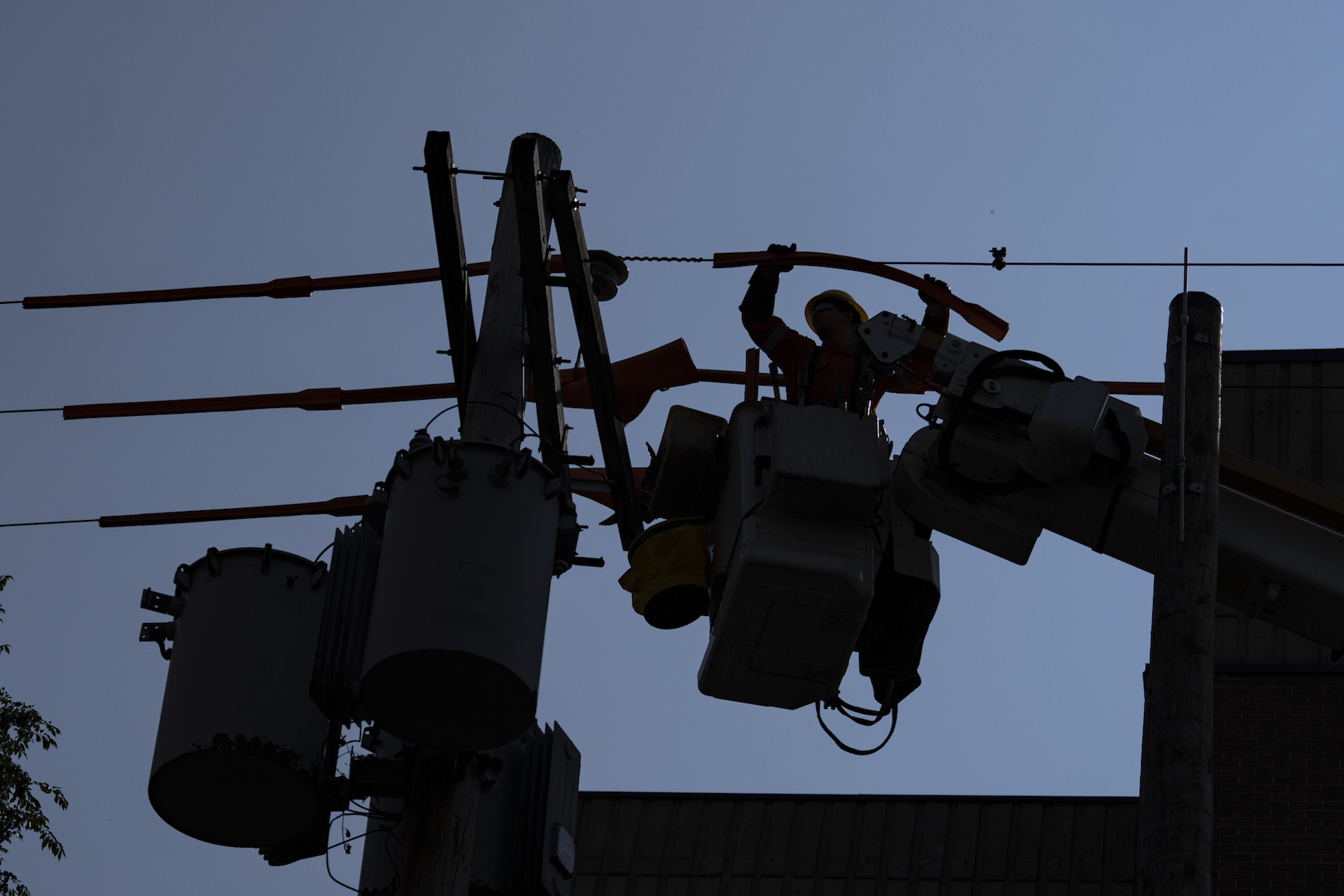 A silhouetted image of a hydro line worker repairing electrical equipment near the top of a utility pole from the bucket of a cherry-picker hoist.
