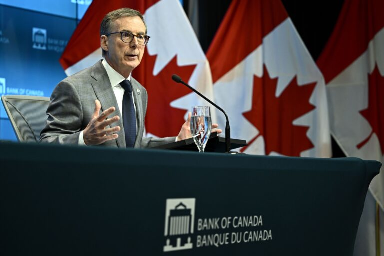 Bank of Canada governor Tiff Macklem speaking into a microphone at a press conference while sitting at a table with the words "Bank of Canada/Banque du Canada" on the tablecloth. Three red-and-white Canadian flags are seen behind him.