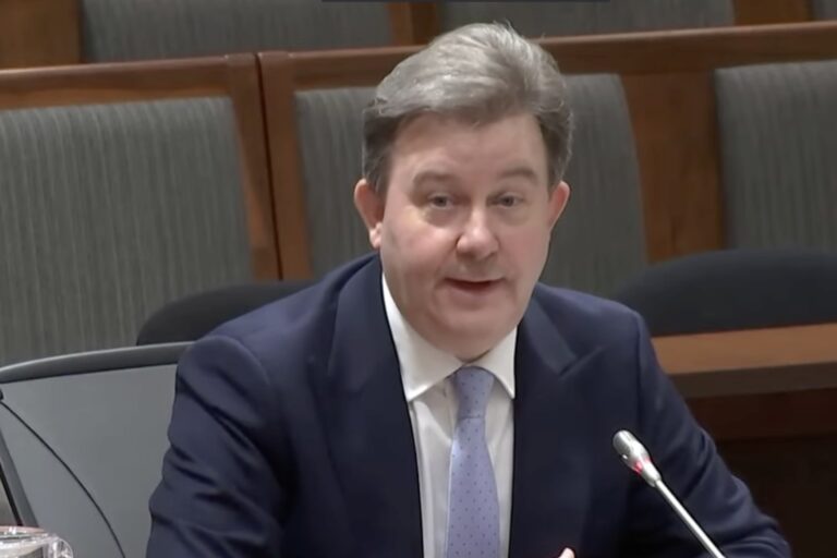 OSFI chief Peter Routledge, in a suit and tie, speaking into a microphone in front of him. There are empty seats in the background.