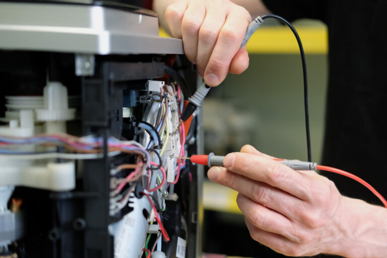 A close-up shot showing the hands of a technician, who is testing internal components of an espresso machine. The person is holding a pen-like electronic sensor in each hand.