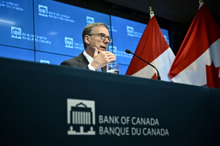 Tiff Macklem, governor of the Bank of Canada, sitting at a desk with the central bank’s logo and speaking into a microphone. There is a big screen and two Canadian flags in the background.