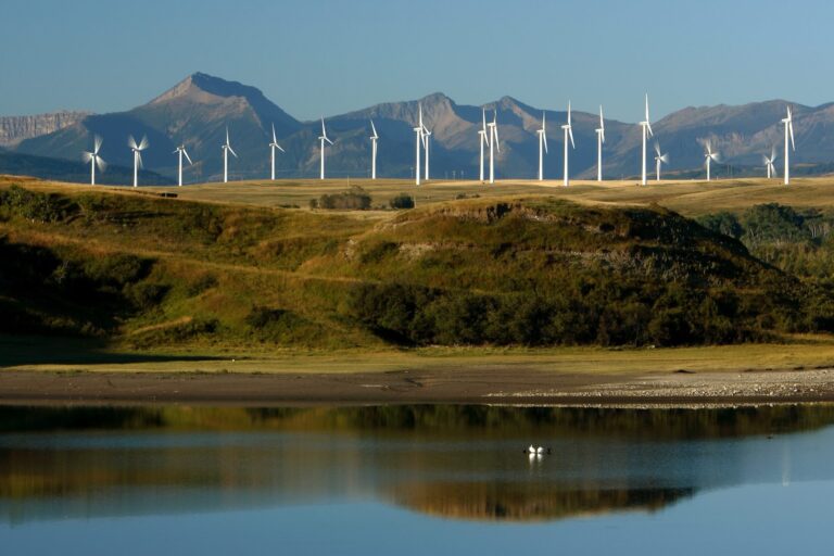 A wind farm with a bunch of windmills on a hill. In the foreground is the Oldman River and in the distance the Rocky Mountains.