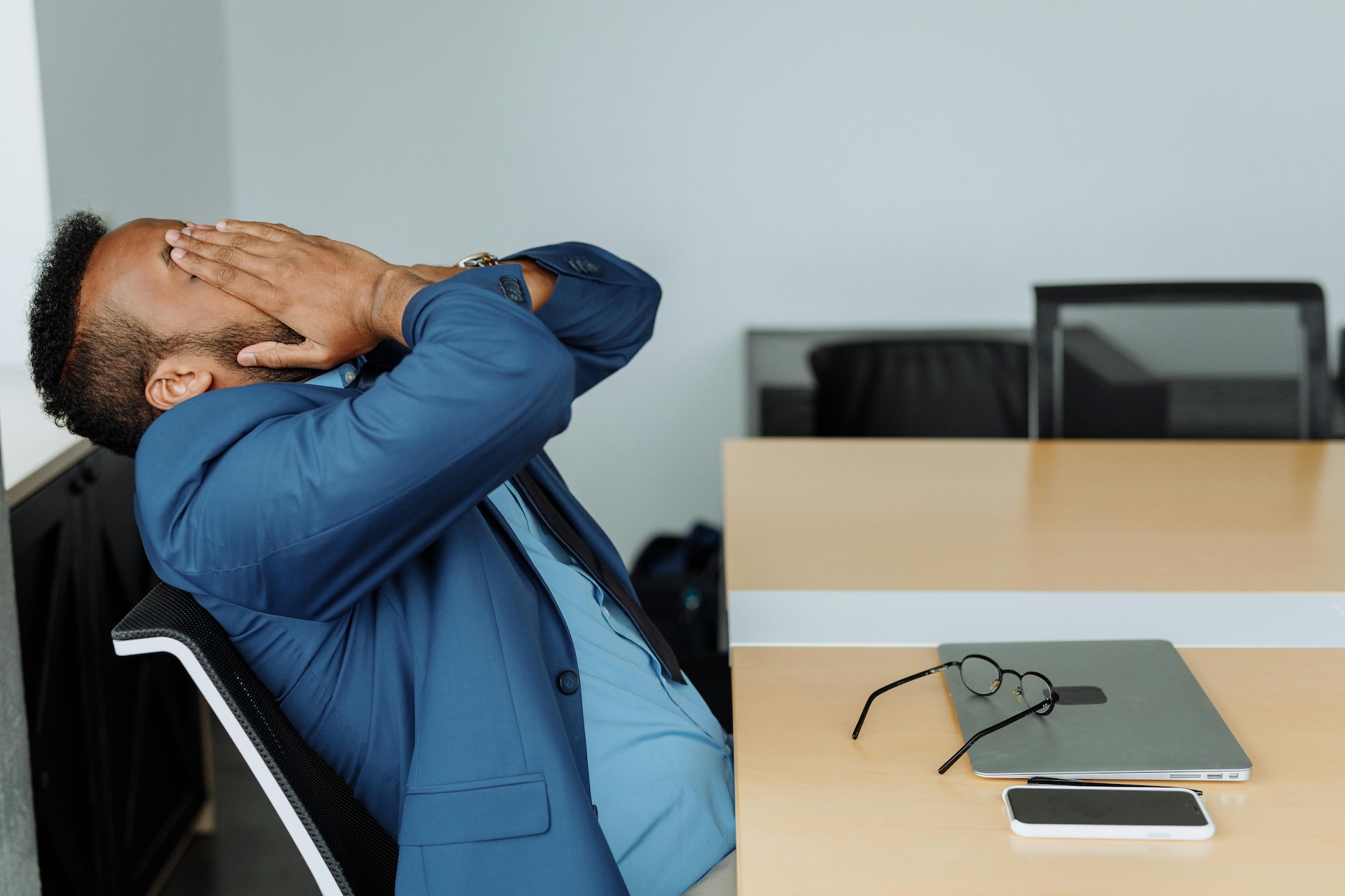 A man in a suit seated at a blond wood table with laptop on it is throwing back his head and holding his hands over his face.
