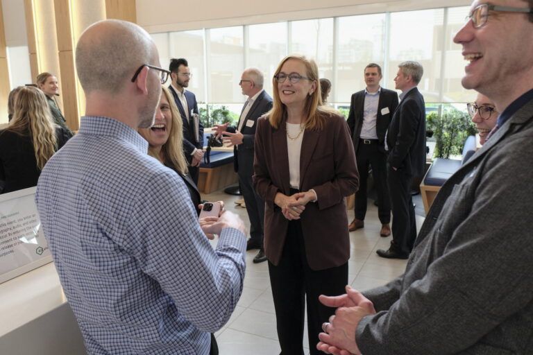 Bailey stands chatting with a small group of people in an airy room before a large bank of windows. She's wearing a burgundy blazer and spectacles with light blue in the rims, and has her hands folded in front of her.