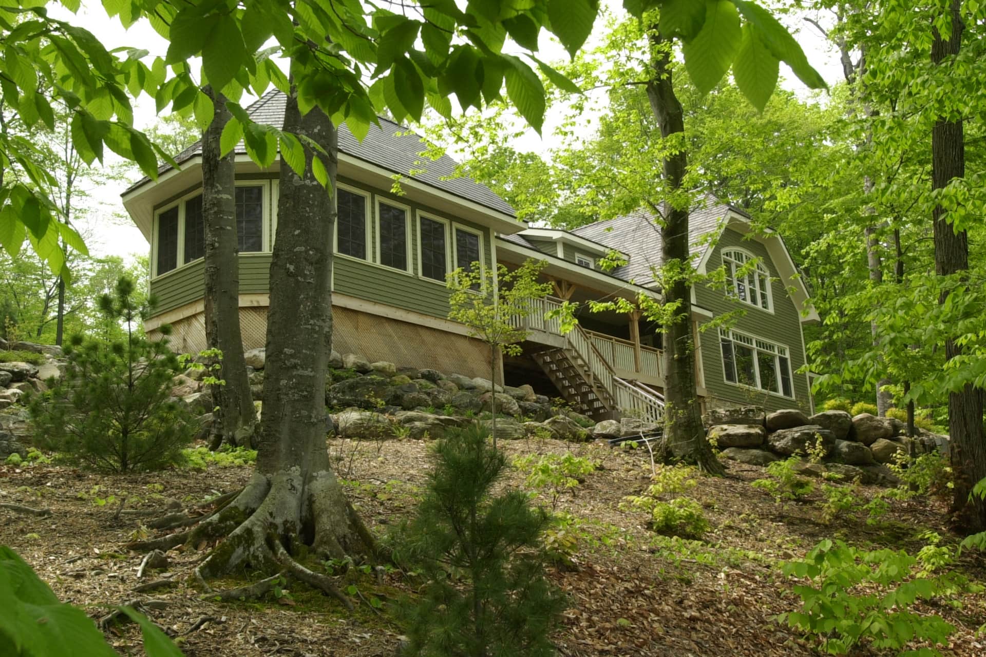 A classic Muskoka cottage with green clapboard, white trim and screened in verandah stands nestled amid leafy trees on a hillside.