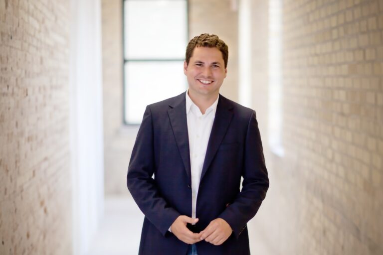 D2L founder John Baker in a dark suit jacket and white shirt. He is standing in a hallway with brick walls and a window in the background, and smiling.