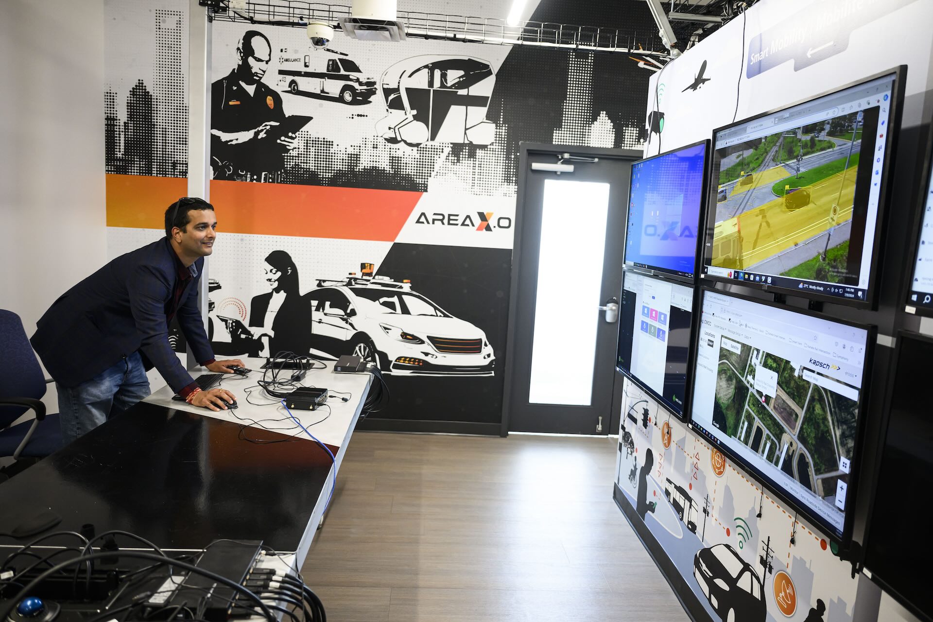 Divyanshu Kamboj of Area X.O at a desk with multiple computer monitors displaying various screens, including maps and aerial views, in a tech control room decorated with automotive and drone graphics.