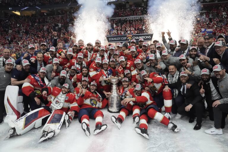 A hockey team poses with the Stanley Cup trophy on an ice rink, surrounded by cheering crowd and celebratory pyrotechnics. Players are wearing red uniforms and championship hats.