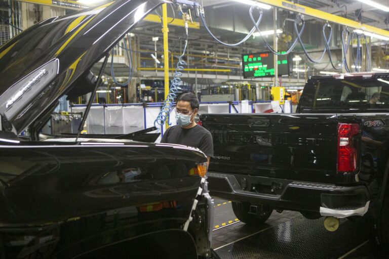 A worker is seen working on an auto assembly line. There are two black 4x4 Chevrolet trucks in front of and behind him.