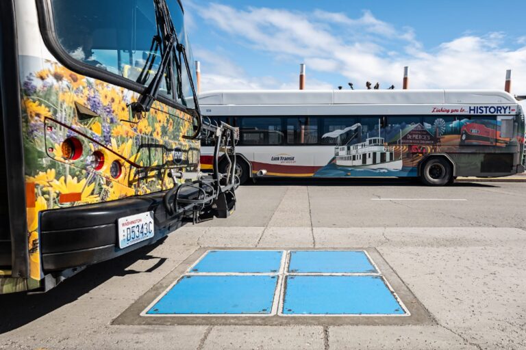 A colourful bus with a Washington number plate parked in a parking lot next to another bus. There are four sky-blue panels grouped together in a square on the ground.