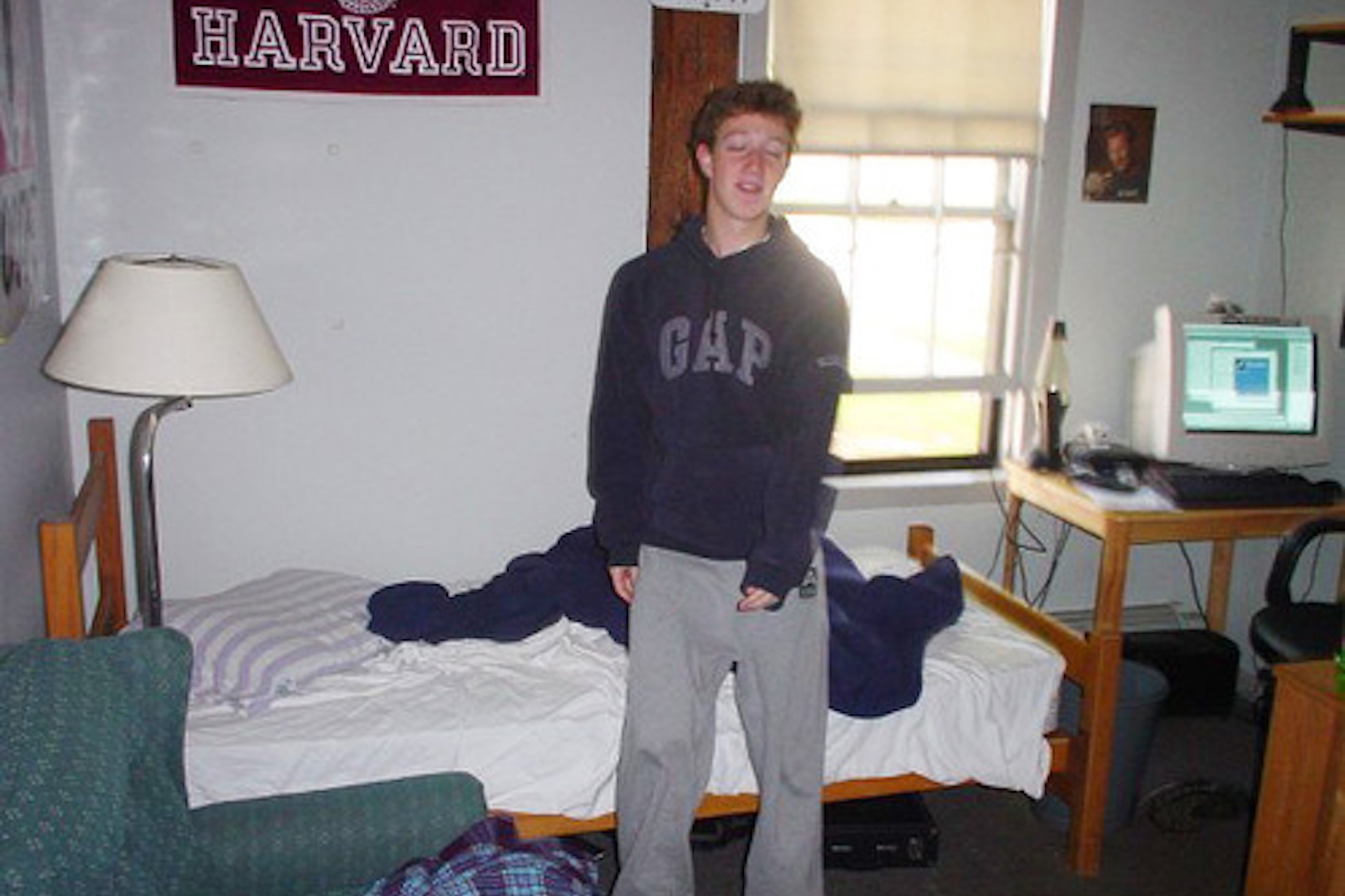 Young Mark Zuckerberg, founder of Facebook, in his dorm room wearing PJs. A sign in the background reads 'HARVARD' in red.